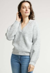 Pull en tricot gris clair avec un col en V et une texture côtelée. Présente des manches longues légèrement bouffantes et une coupe décontractée.