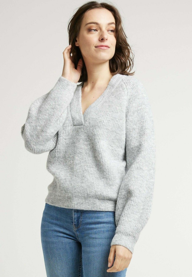 Pull en tricot gris clair avec un col en V et une texture côtelée. Présente des manches longues légèrement bouffantes et une coupe décontractée.