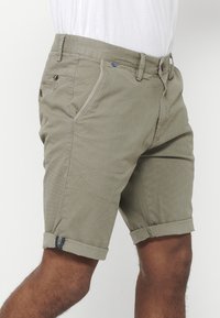 Beige textuuroverhemd met opgerolde manchetten, gedragen door een persoon tegen een effen witte achtergrond.