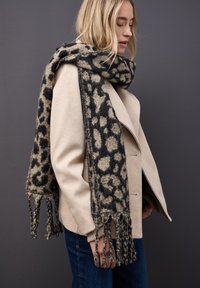 Sjal med leopardmønster i blødt stof, med sorte og beige farver, frynser og lagt over en lys farvet frakke.
