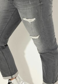 Jean en denim gris avec une finition délavée, présentant deux déchirures au niveau des genoux et des ourlets retroussés, accompagné de bottines argentées brillantes.