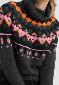 Šedý pletený svetr s vysokým límcem, který má geometrický vzor v oranžové, růžové a zelené barvě na hrudi a rukávech.