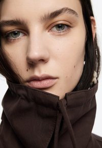 Zbliżenie na twarz w brązowym ubraniu z wysokim kołnierzem. Cechy: czysta skóra, jasne brwi i neutralny kolor ust.