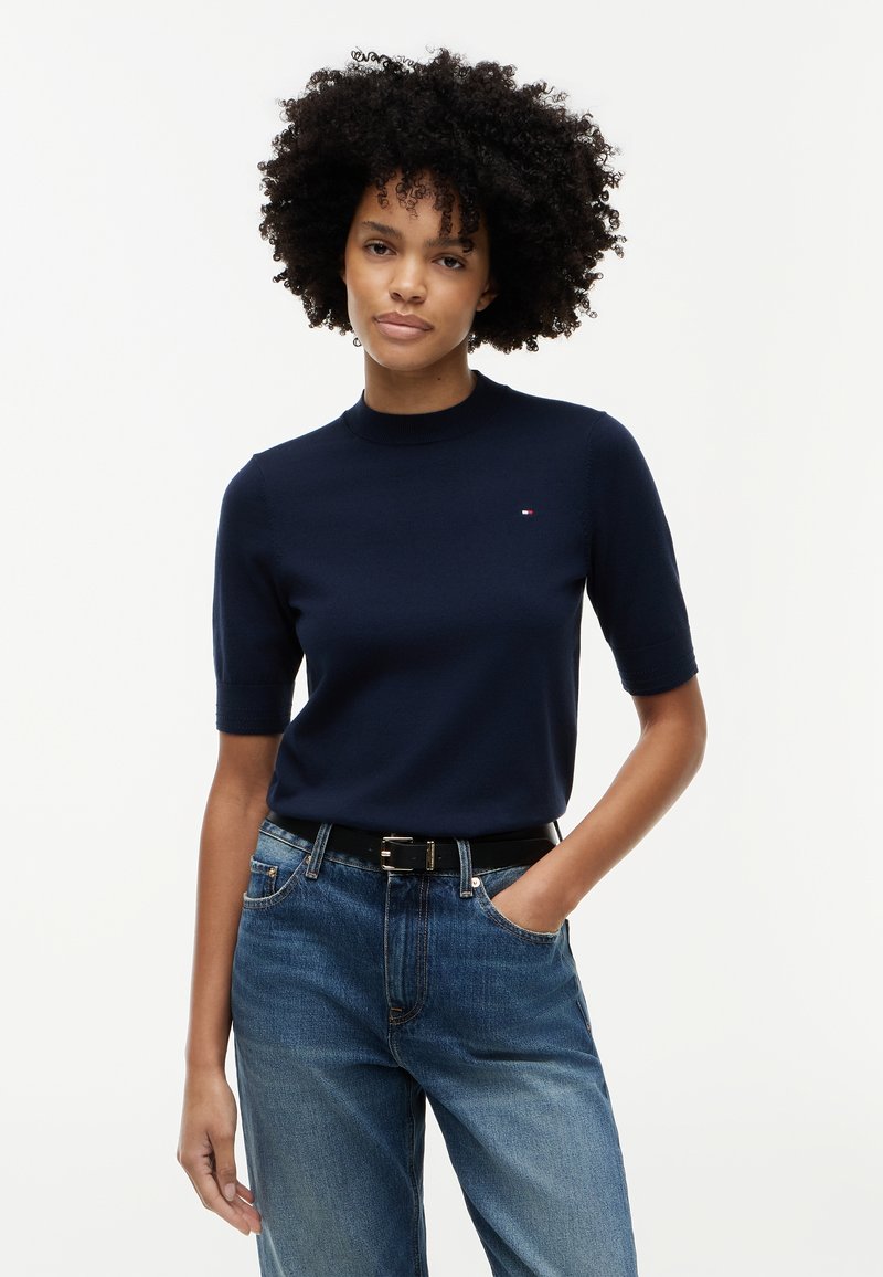 Femme aux cheveux bouclés portant un haut bleu marine à manches courtes et un jean bleu, debout avec une main dans la poche devant un fond uni.