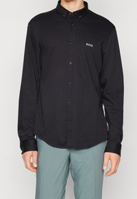 Schwarzes Langarmhemd mit Knopfleiste am Kragen und dezentem Logo auf der Brust. Verfügt über kontrastierende Manschetten und eine glatte Stoffstruktur.