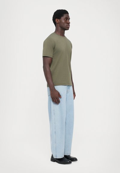 Oliven grøn t-shirt med korte ærmer, afslappet pasform, parret med lyseblå vide jeans og sorte sko, stående på en hvid baggrund.