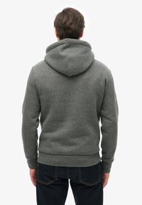 Sudadera con capucha gris, confeccionada con tela suave, que presenta un diseño limpio, puños y dobladillo acanalados, con un bolsillo tipo canguro en la parte frontal.