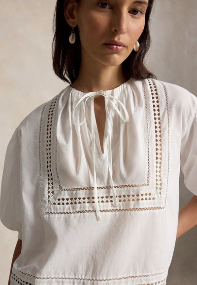 Γυναίκα που φοράει λευκό μπλουζάκι με δαντελένιες λεπτομέρειες και τετράγωνη κοπή, με δέσιμο στον λαιμό, συνοδευόμενο από σκουλαρίκια σε σχήμα κοχυλιού, ουδέτερο φόντο.