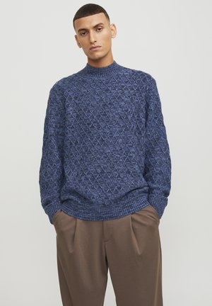 Suéter de punto en azul oscuro con un patrón de diamantes texturizado, con cuello alto y puños acanalados, combinado con pantalones marrones de corte holgado.