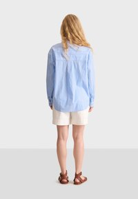 Camicia azzurro chiaro a maniche lunghe con righe bianche verticali, dotata di una piega sul retro. Abbinata a pantaloni corti color crema e sandali marroni.