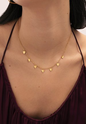 Zlatý náhrdelník s reťazou so siedmimi malými príveskami v tvare srdca, ktorý nosí žena na krku cez bordový top na tenkých ramienkach.