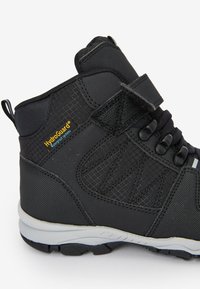 Schwarze wasserdichte Stiefel mit strukturierter Oberseite, ausgestattet mit einem Klettverschluss, elastischen Schnürsenkeln und einer grauen Gummisohle. HydroGuard® Branding ist sichtbar.