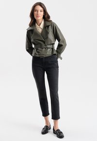 Olijfgroene neplederen jas met een brede kraag, een ceintuur in de taille en zijkleppen, gecombineerd met zwarte slim-fit jeans en loafers.