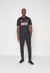 Camiseta deportiva negra con el logo rosa de "MESSI", combinada con pantalones ajustados a juego y zapatillas deportivas negras. Diseño simple y deportivo con branding.