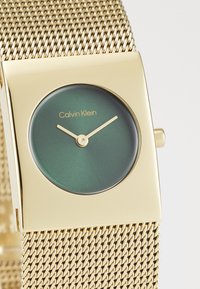 Calvin Klein watch with a round green dial, gold hour and minute hands, and a gold mesh bracelet. Square case design with minimalist styling.