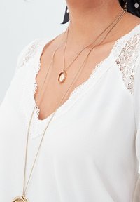 Witte top met kantafwerking bij de halslijn, met delicate details. Twee lagen gouden kettingen, waarvan er één een textured hanger heeft.
