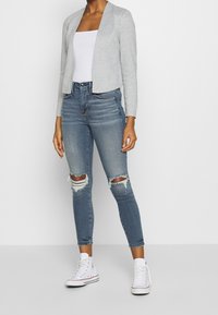 Harmaa, puolipitkä neuletakki avonaisella edellä, yhdistettyään revittyihin sinisiin skinny-farkkuihin ja valkoisiin lenkkareihin. Sileä kangas ja rento istuvuus.