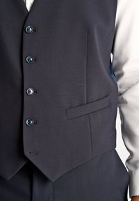 Dunkelblauer Weste mit fünf Knöpfen, Reverskragen und einer einzelnen Tasche. Glatter, taillierter Stoff und scharfe Kanten, kombiniert mit einem weißen Hemd.
