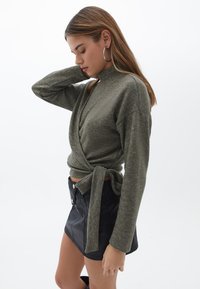Oliwkowy sweter z długim rękawem o kroju kopertowym i przednim wiązaniu. Połączony z czarną, błyszczącą mini spódnicą z guzikami i kieszeniami.