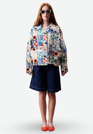 Femme portant une veste à motifs surdimensionnée, un short en jean foncé, des ballerines rouges tissées et de grandes lunettes de soleil à monture blanche, debout devant un fond blanc.