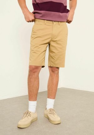 Person trägt beige knielange Shorts, beige Turnschuhe, weiße Socken und ein bordeaux gestreiftes Hemd und steht auf einem grauen Boden.