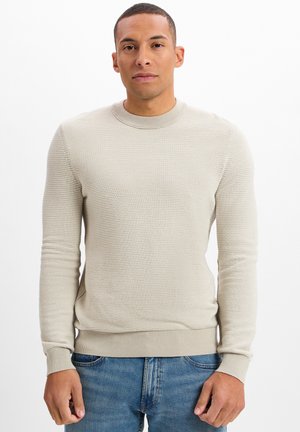 Beige Ribstrickpullover mit Rundhalsausschnitt und langen Ärmeln, mit einer enganliegenden Saumkante und Bündchen. Kombiniert mit blauen Denim-Jeans.
