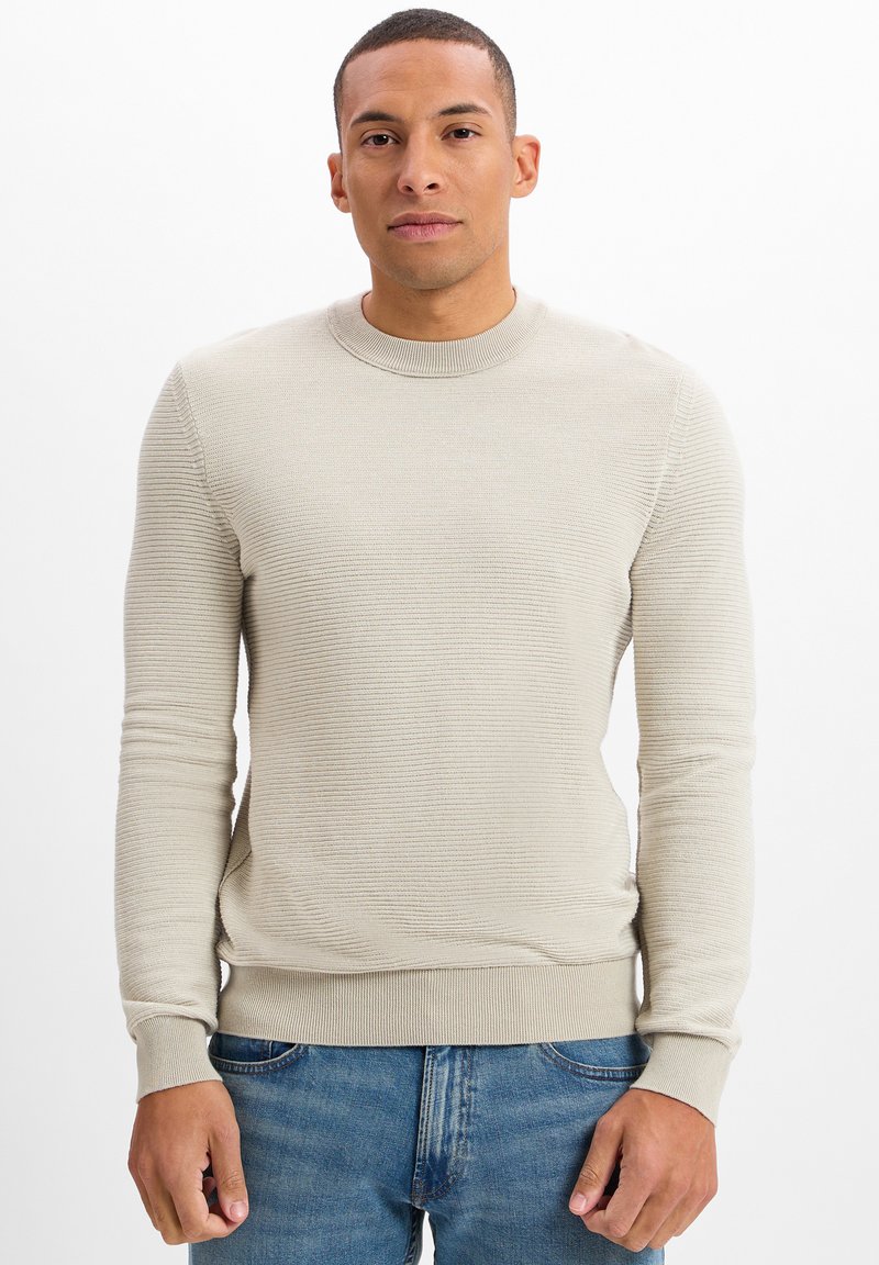 Beige geribbelde gebreide trui met ronde hals en lange mouwen, met een aansluitende onderkant en manchetten. Gecombineerd met blauwe denim jeans.