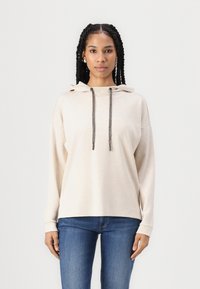 Hoodie bege com cordão e cordões cinzentos contrastantes, apresentando um corte relaxado e mangas longas, combinado com jeans de ganga escura.