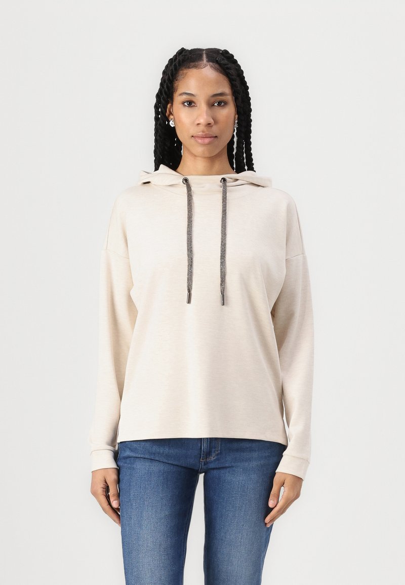 Hoodie bege com cordão e cordões cinzentos contrastantes, apresentando um corte relaxado e mangas longas, combinado com jeans de ganga escura.