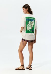 Cream-colored oversized t-shirt with a green and blue shell graphic on the back. Paired with striped shorts and brown strappy sandals.