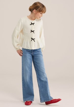 Joven que mira hacia abajo, vestida con una blusa texturizada color crema con lazos negros, jeans azules de pierna ancha y zapatos planos rojos perforados, sobre un fondo liso.