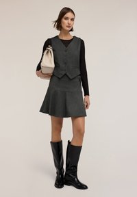 Graue Weste mit enganliegendem Design, kombiniert mit einem grauen, ausladenden Rock. Das Model trägt glänzende schwarze knielange Stiefel und hält eine hellbeige Handtasche.