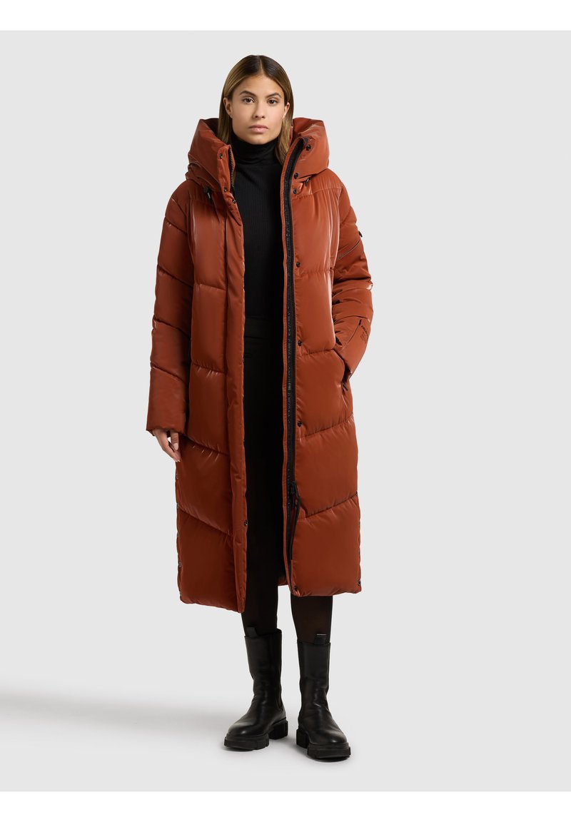 Abrigo largo acolchado en naranja óxido, con cuello alto, cierre de cremallera y diseño acolchado. Lleva puesto sobre un conjunto negro y botines hasta el tobillo.