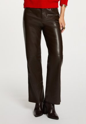 Donna vestita con pantaloni di pelle marrone scuro, stivali alla caviglia in pelle abbinati, maglione rosso, bracciale dorato e anello, in piedi davanti a uno sfondo neutro.
