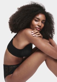 Frau mit lockigem Haar sitzt, trägt schwarzen Spitzen-BH und passende Unterwäsche, legt die Arme auf die Knie auf weißem Hintergrund.