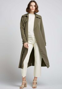 Manteau long vert olive à l'avant ouvert, associé à un pull crème et un pantalon léger court. Des sandales beige à talon épais complètent le look.