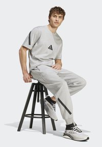 Šedé bavlněné tričko s černými akcentními pruhy, v kombinaci se šedými tepláky odpovídající barvy, které mají boční kapsy a černé detaily. Černobílé tenisky.