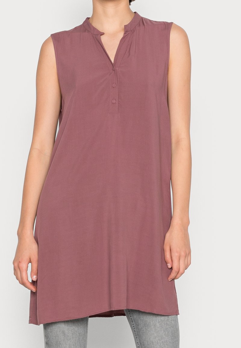 Tunique mauve sans manches avec une courte patte de boutonnage portée sur un pantalon gris sur une personne devant un fond clair.