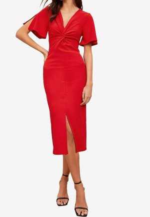 Mujer que lleva un vestido midi rojo con mangas con vuelo, un detalle de nudo delantero y una abertura frontal, combinado con tacones negros con pulsera en el tobillo.