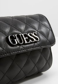 Czarna pikowana torebka z błyszczącego materiału z logo "GUESS". Tekstura jest gładka, z wypukłymi wzorami w diamenty i usztywnioną konstrukcją.