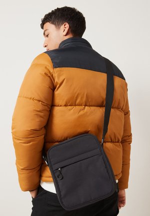 Uomo con giacca imbottita nera e marrone e una borsa nera a tracolla su uno sfondo chiaro e uniforme.