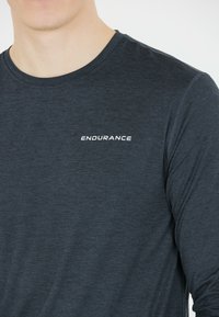 Maglietta sportiva a maniche lunghe in tessuto grigio scuro con una leggera trama. Presenta il logo bianco "ENDURANCE" sul petto.