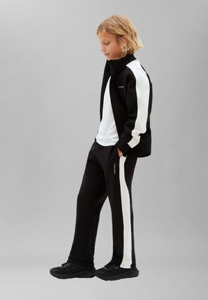 Blond laps seisab käed taskutes, seljas musta ja valget toonides spordijakk ja püksid ning mustad tossud, halli taustaga.