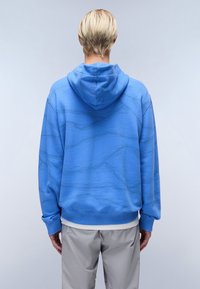 Hanorac albastru cu glugă, având un design texturat cu linii ondulate, manșete ribbate și o croială lejeră. Se poartă pantaloni gri dedesubt.
