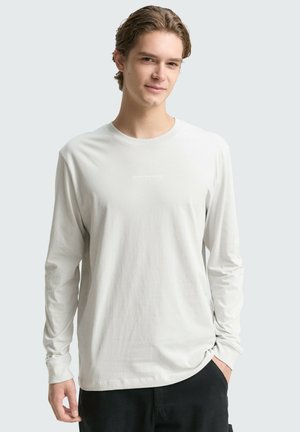 Młody mężczyzna z jasnobrązowymi włosami, ubrany w prostą białą koszulę z długim rękawem i czarne spodnie, stoi z jedną ręką w kieszeni.