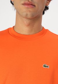 Tricou din bumbac portocaliu cu gât rotund și mâneci scurte, având un mic logo verde de crocodil pe partea stângă a pieptului.