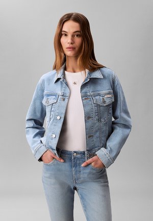 Donna con giacca di jeans azzurro chiaro e jeans, con sotto una maglietta bianca a coste, in piedi con le mani nelle tasche contro uno sfondo grigio.