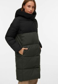 Paminkštintas paltas su juoda viršutine dalimi ir tamsiai žalia apatine dalimi, turintis aukštą apykaklę, du šoninius kišenes ir kviltuotą tekstūrą.