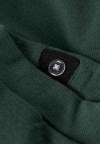 Zaļš kokvilnas audums ar melnu etiķeti, pie kuras pievienots caurspīdīgs, apaļš pogs, akcentējot tekstūru un šuvju detaļas.