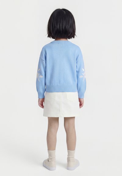 Kind met kort zwart haar staand, draagt een lichtblauwe trui met witte bloemmotieven, een witte rok, beige sokken en sneakers.